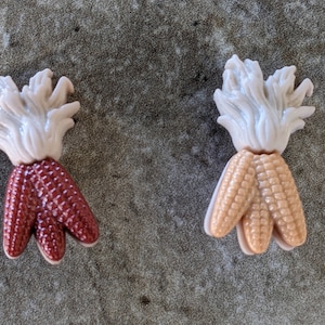 May include: Two plastic corn on the cob decorations. One is red and the other is yellow.