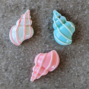 May include: Three seashell-shaped decorative items. One is white with pink stripes, another is light blue with white stripes, and the third is pink with white stripes. The items are on a textured gray surface.