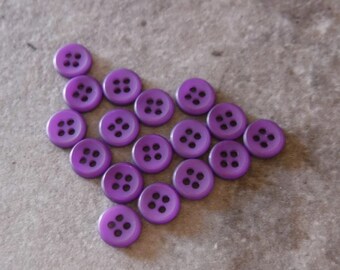 May include: A collection of 14 purple plastic buttons with four holes each. The buttons are arranged in a triangular shape.