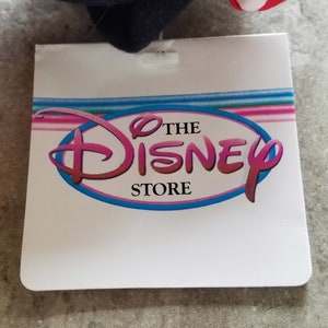 Może przedstawiać: Biała metka z niebieskim owalem zawierającym tekst "The Disney Store" w r&oacute;żowych literach.