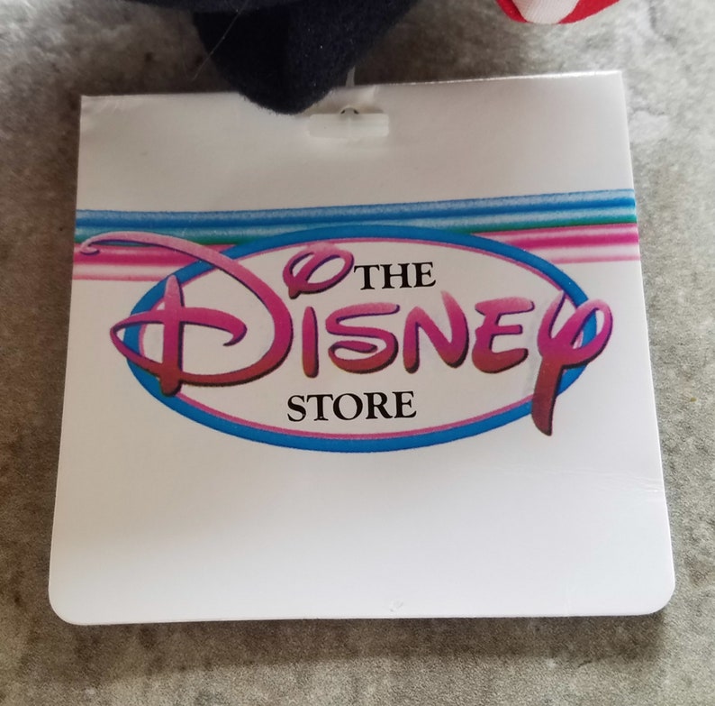 Może przedstawiać: Biała metka z niebieskim owalem zawierającym tekst "The Disney Store" w r&oacute;żowych literach.