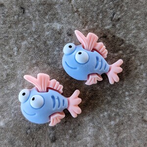 May include: Two blue and pink fish-shaped charms with white eyes and a smiling mouth. The charms are made of a soft, pliable material.
