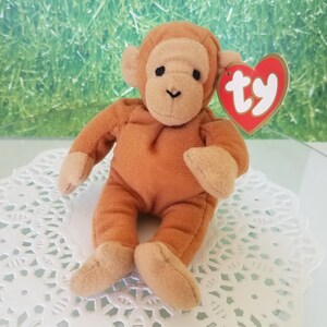 May include: A brown plush monkey toy with a Ty tag that says "ty" in red letters. The monkey is sitting on a white doily.