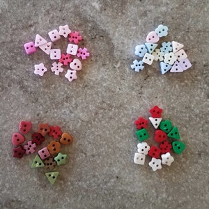 May include: Assortment of colorful plastic buttons in various shapes, including hearts, stars, leaves, and triangles. The buttons are arranged in four groups on a gray surface.