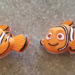 May include: Two orange and white clownfish figurines with black outlines and large, round eyes. The figurines are made of plastic and have a 3D design.