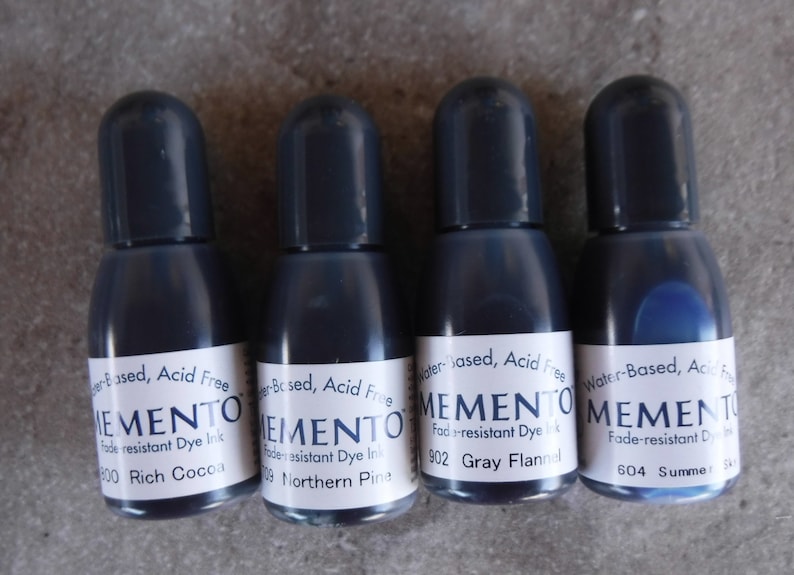 Puede incluir: Cuatro botellas peque&ntilde;as de tinta de tinte resistente a la decoloraci&oacute;n Memento. Las botellas son de color azul oscuro con tapas a juego. Cada botella tiene una etiqueta blanca con el nombre de la marca y el color de la tinta.