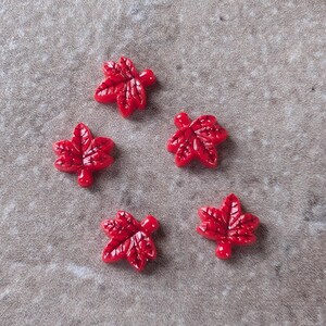 May include: Five small, glossy red buttons shaped like maple leaves. Each button features a detailed, embossed vein pattern on the surface, capturing the distinct five-pointed structure of a leaf. The buttons have a smooth, reflective finish.