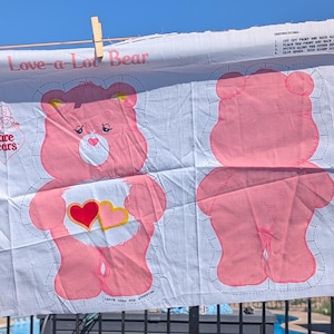 May include: A white fabric panel featuring Care Bears characters, including Grumpy Bear and Love-a-Lot Bear. The bears are in shades of blue and pink, with heart and cloud details. The words "Care Bears" and bear names are printed on the fabric.