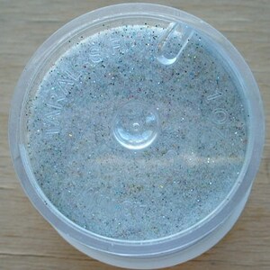 May include: A small, clear plastic container filled with iridescent glitter. The glitter is a mix of silver, white, and light blue.