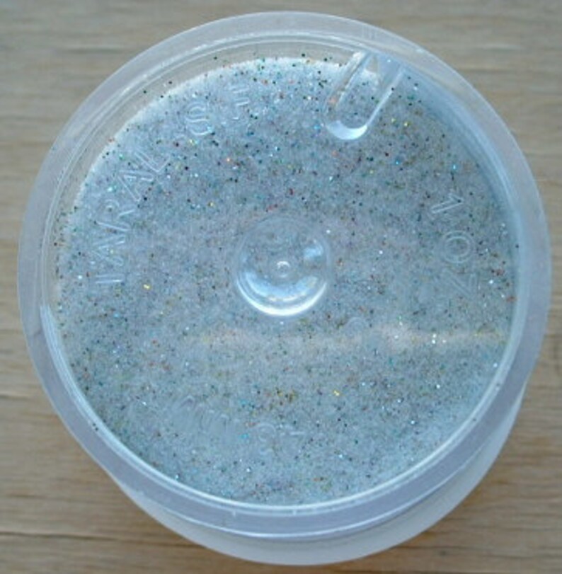 May include: A small, clear plastic container filled with iridescent glitter. The glitter is a mix of silver, white, and light blue.