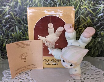 May include: A porcelain figurine of a smiling child hanging upside down with a string attached to their foot. The figurine is wearing a light blue and white patterned outfit and has a light green hat. The figurine is part of a Precious Moments collection and the box reads "Smile Along The Way".