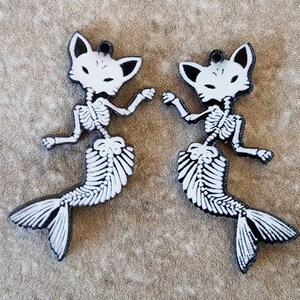 May include: Two white skeleton mermaid charms with a black outline. The mermaids have a fox-like head and a fish tail.