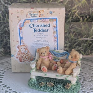 May include: A figurine of two teddy bears sitting on a white bench with a blue banner that reads "Heart to Heart." The bears are light brown with pink and blue ribbons. The box behind the bears reads "Cherished Teddies."