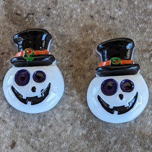 May include: Two white ceramic Halloween pumpkin ornaments with black eyes, a black mouth, and a black top hat with an orange and green band.