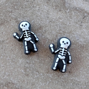 May include: Two black and white skeleton charms. Each charm features a black body with a white skeleton design, including a skull head and rib cage. The charms are small and appear to be made of a rubbery material.