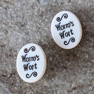 May include: Two white round ceramic ornaments with black lettering and decorative swirls. The text on the ornaments reads "Worm's Wort".