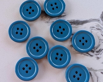 May include: A collection of nine teal-colored buttons with four holes each. The buttons are round and have a slightly raised edge. They are arranged on a light gray and white marbled surface.