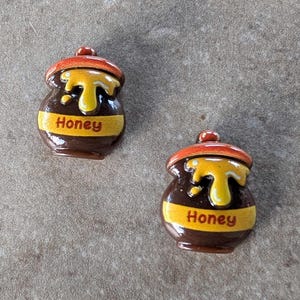 May include: Two ceramic honey pots with brown bodies, orange lids, and yellow honey dripping down the sides. Each pot has a yellow band with the word "Honey" in red. The pots are on a textured, gray surface.