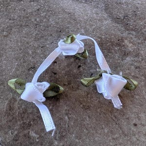 May include: Two white ribbon roses with green leaves. Each rose is made of folded white ribbon and has a small, tightly wound center. The green leaves are oval-shaped and add a touch of color. These delicate floral embellishments are ideal for crafts.