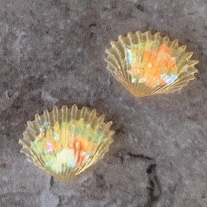 May include: Two translucent, seashell-shaped objects with iridescent interiors. The shells have a ridged, fan-like design and display a spectrum of colors, including orange, yellow, and green. They rest on a mottled gray surface.