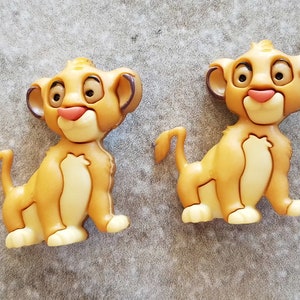 May include: Two Simba-shaped buttons. The buttons are light brown with a cream-colored belly and paws. They have large, expressive eyes, a pink nose, and a small tail. The buttons are made of a glossy material and are set against a gray background.