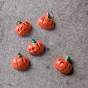 May include: Five small, glossy, orange pumpkin-shaped cabochons with green stems and dark orange vertical indentations. Each pumpkin features a smooth, rounded surface and a distinct, raised green stem at the top, arranged in a scattered pattern.