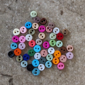 May include: A collection of colorful plastic buttons in various shades of pink, blue, green, orange, brown, and white. The buttons are small and have two holes each.