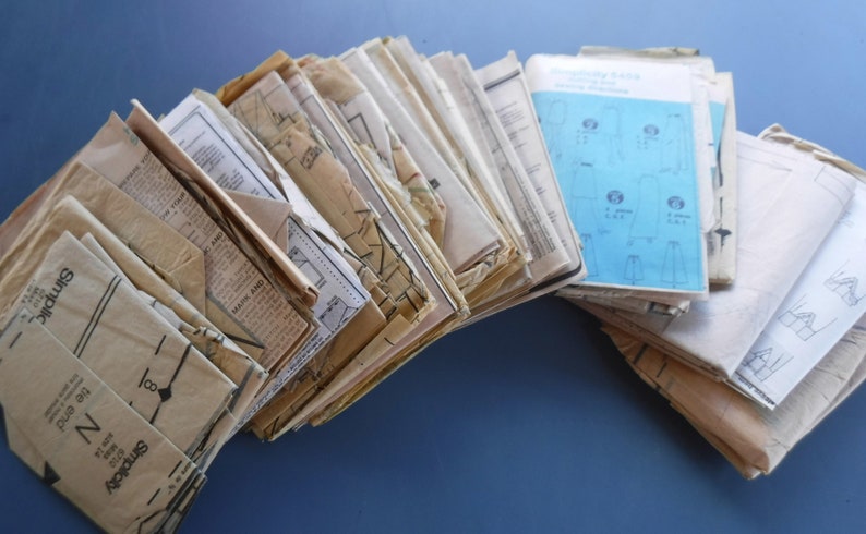 May include: A stack of vintage sewing patterns in various colors and sizes. The patterns are mostly brown and white with some blue and green accents. The patterns are folded and stacked on top of each other.