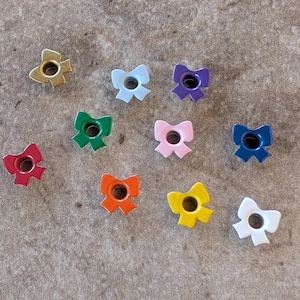 May include: A collection of colorful bow-shaped metal embellishments in various colors, including gold, red, green, orange, yellow, pink, blue, purple, and white. Each bow has a central hole, suitable for crafting or decorative purposes.