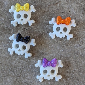May include: Four white skull-shaped charms with glitter bows. The bows are yellow, orange, black, and purple.