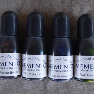 Puede incluir: Cuatro peque&ntilde;os frascos de tinta con tapas de color gris oscuro. Cada frasco tiene una etiqueta blanca con la palabra "MEMENTO" y el nombre del color de la tinta. Los colores incluyen Peanut Brittle, Pistachio, Danube Blue y Pear Tart. La tinta es a base de agua y resistente a la decoloraci&oacute;n.