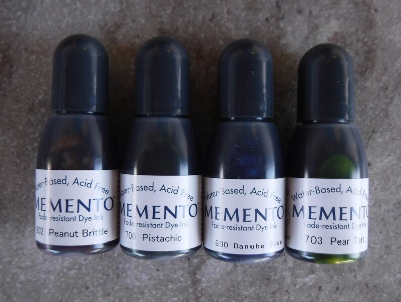 Puede incluir: Cuatro peque&ntilde;os frascos de tinta con tapas de color gris oscuro. Cada frasco tiene una etiqueta blanca con la palabra "MEMENTO" y el nombre del color de la tinta. Los colores incluyen Peanut Brittle, Pistachio, Danube Blue y Pear Tart. La tinta es a base de agua y resistente a la decoloraci&oacute;n.