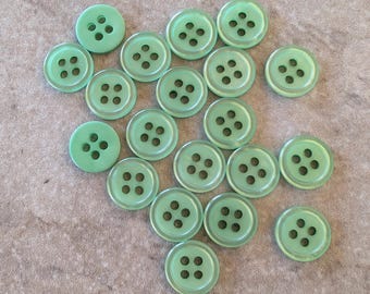 May include: A collection of light green, round buttons with four holes each. The buttons are scattered on a textured, neutral-toned surface. The buttons are made of a translucent material.