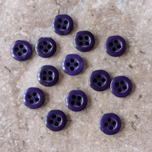 May include: A collection of fourteen dark purple buttons with four holes each. The buttons are round and have a glossy finish. They are scattered on a light brown, textured surface.