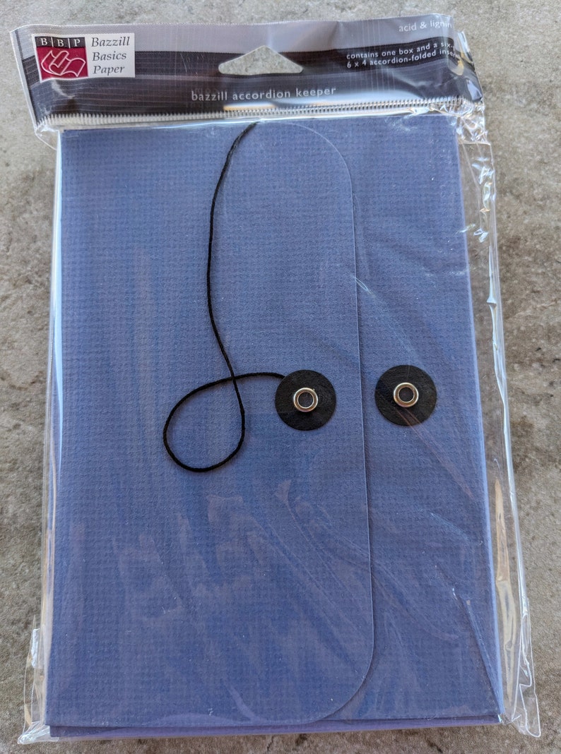 May include: A blue accordion keeper made of paper with a black string and two metal eyelets. The packaging says "Bazzill Basics Paper" and "bazzill accordion keeper".