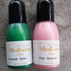 Op de afbeelding: Twee kleine flesjes Delicata Inker, &eacute;&eacute;n smaragdgroen en &eacute;&eacute;n roze met shimmer. Beide flesjes zijn gelabeld met de merknaam en kleur, en hebben een inhoud van 0,5 fl.oz (15ml).