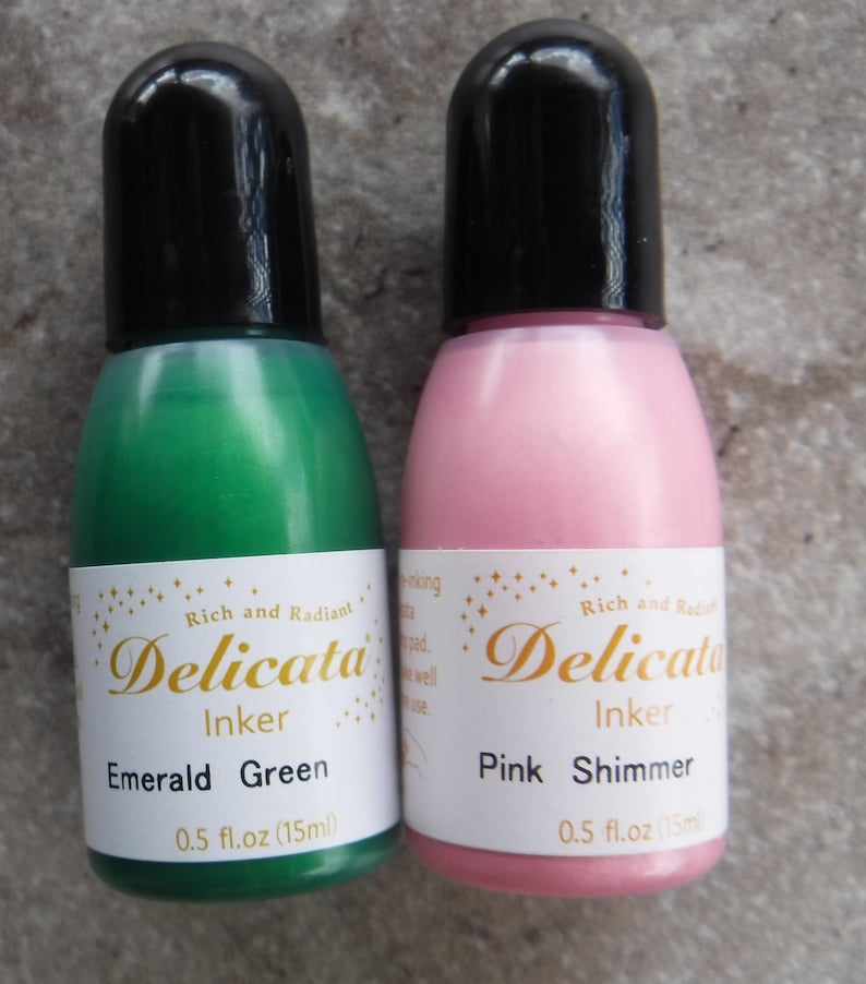 Op de afbeelding: Twee kleine flesjes Delicata Inker, &eacute;&eacute;n smaragdgroen en &eacute;&eacute;n roze met shimmer. Beide flesjes zijn gelabeld met de merknaam en kleur, en hebben een inhoud van 0,5 fl.oz (15ml).