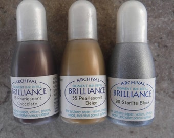 May include: Three bottles of Archival Brilliance pigment ink refill in Pearlescent Chocolate, Pearlescent Beige, and Starlite Black. The bottles are labeled with the ink color and the brand name.