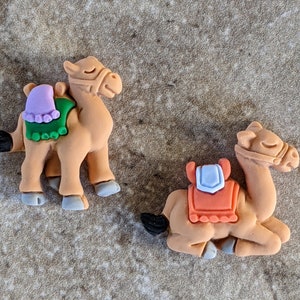May include: Two brown camel figurines with saddles. The camel on the left is standing with a green and purple saddle. The camel on the right is lying down with an orange and white saddle.