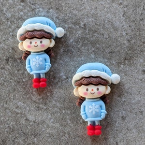 May include: Two blue and white Christmas ornaments shaped like girls wearing blue sweaters with snowflakes and red boots. The girls have brown hair and brown eyes.