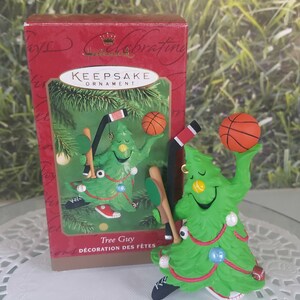 May include: A green Christmas tree ornament shaped like a tree with a smiling face. The tree is wearing sneakers and holding a basketball, baseball bat, hockey stick, and soccer ball. The ornament is labeled "Tree Guy" and "Décoration des Fêtes".