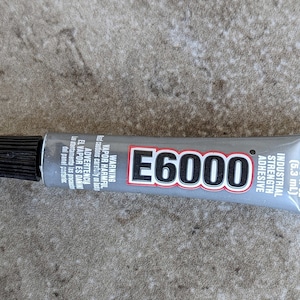 May include: A gray E6000 industrial strength adhesive tube with a black cap. The label features the text E6000, Industrial Strength Adhesive, 0.18 fl oz (5.3 mL), and warning text in English and Spanish regarding harmful vapors.
