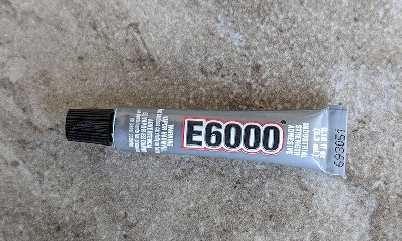May include: A gray E6000 industrial strength adhesive tube with a black cap. The label features the text E6000, Industrial Strength Adhesive, 0.18 fl oz (5.3 mL), and warning text in English and Spanish regarding harmful vapors.