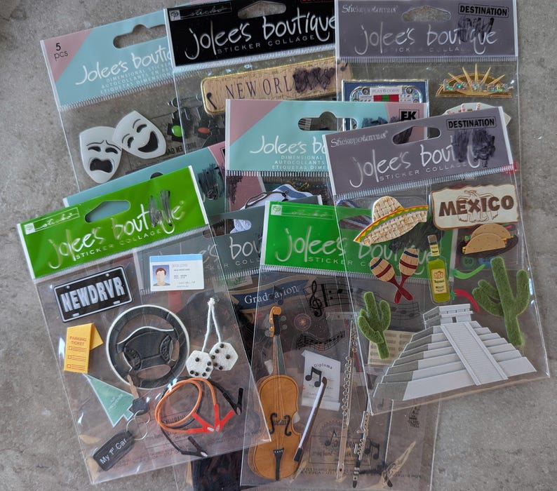 May include: A collection of Jolee's Boutique sticker collages. The stickers feature various themes, including travel, music, and driving. Some stickers have text like "NEWDRVR", "MEXICO", and "Graduation". The stickers are in clear plastic packaging.