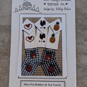 May include: A pattern booklet titled "The Village Peddler" featuring designs by Kathy Nobis. The booklet showcases images of pot holders and tea towels with heart, rooster, and pineapple motifs. The pot holders and tea towels are in a plaid pattern.