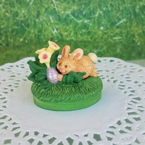 May include: A small ceramic figurine of a brown bunny rabbit holding a purple Easter egg, surrounded by green leaves and yellow flowers.