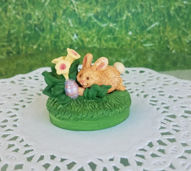 May include: A small ceramic figurine of a brown bunny rabbit holding a purple Easter egg, surrounded by green leaves and yellow flowers.
