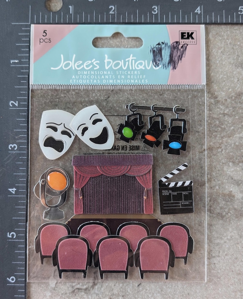 May include: A package of 5 dimensional stickers featuring theater-themed designs. The stickers include comedy and tragedy masks, stage lights, a stage with curtains, a film clapperboard, and rows of theater seats. The package is labeled "Jolee's Boutique".