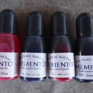 Puede incluir: Cuatro botellas peque&ntilde;as de tinta Memento a base de agua, sin &aacute;cido y resistente a la decoloraci&oacute;n. Colores: Angel Pink, Paris Dusk, Morocco y Elderberry. Cada botella tiene una tapa negra y una etiqueta blanca.