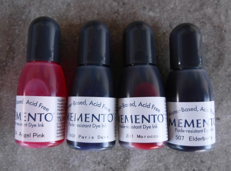 Puede incluir: Cuatro botellas peque&ntilde;as de tinta Memento a base de agua, sin &aacute;cido y resistente a la decoloraci&oacute;n. Colores: Angel Pink, Paris Dusk, Morocco y Elderberry. Cada botella tiene una tapa negra y una etiqueta blanca.